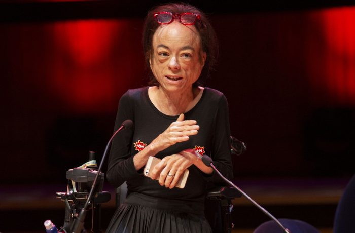 Liz Carr sits behind two microphones in a dark room with red backlighting. She has short hair with red glasses perched on her head, and is clutching a phone.