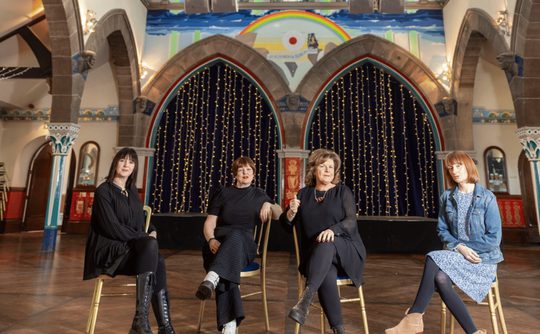 Four women sat on chairs facing the camera, they are in an old church with stone arches. The chairs are arranged at slightly different angles, giving this picture a sense of playfulness.