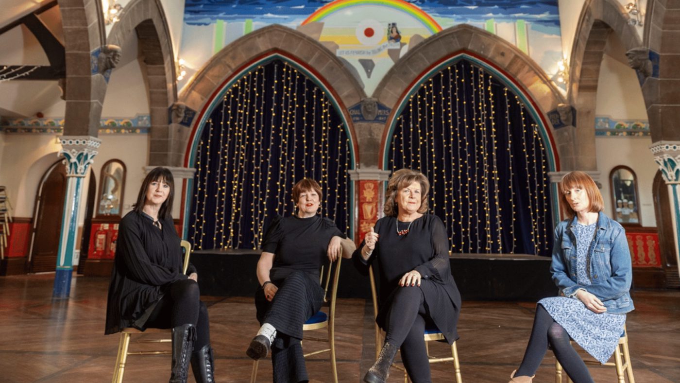 Four women sat on chairs facing the camera, they are in an old church with stone arches. The chairs are arranged at slightly different angles, giving this picture a sense of playfulness.