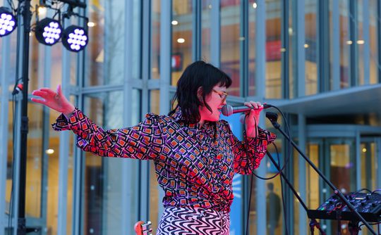 Woman in colourful clothing sings into a microphone, eyes closed, with one arm extended.