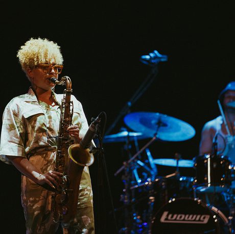 A musician plays the saxophone on stage, wearing sunglasses and a patterned outfit. Behind them, a drummer. The scene is dimly lit
