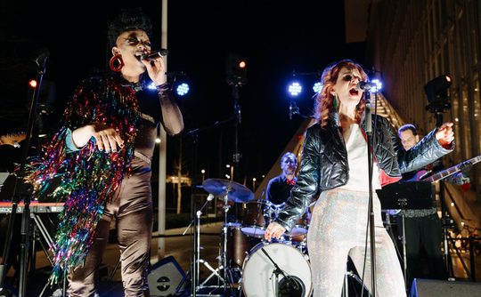 Two singers perform on an outdoor stage with a band behind them. They are wearing elaborate shiny outfits.