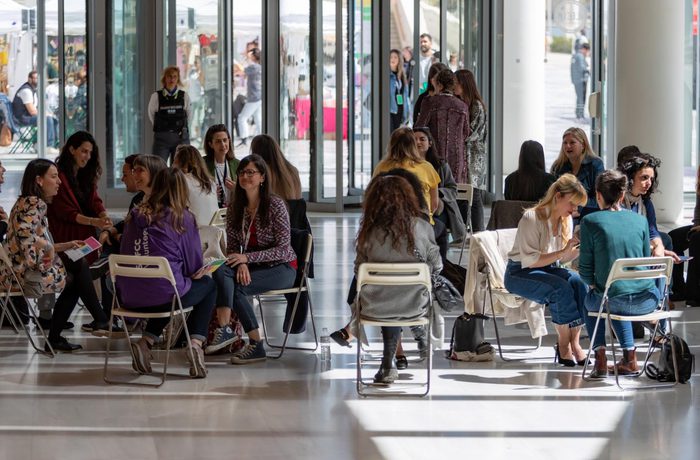 Groups of people sit in folding chairs in a sunlit room with large windows, engaged in lively conversations.