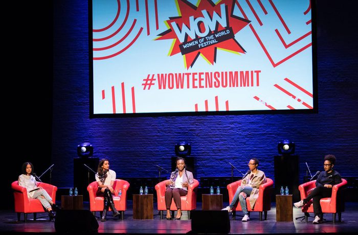 Five women sit on bright red chairs on stage beneath a projected screen reading ‘WOW Women of the World Festival #wowteensummit’. The woman in the centre is speaking and looking at the audience.