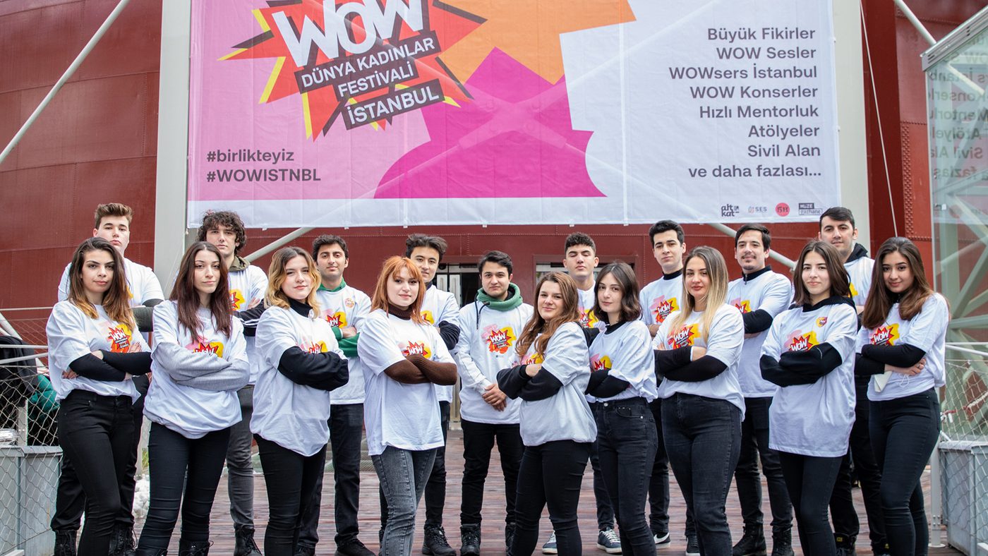 A group of young individuals standing confidently with crossed arms in front of a large, colourful "WOW Dünya Kadınlar Festivali İstanbul" banner