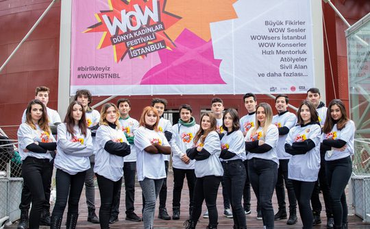 A group of young individuals standing confidently with crossed arms in front of a large, colourful "WOW Dünya Kadınlar Festivali İstanbul" banner