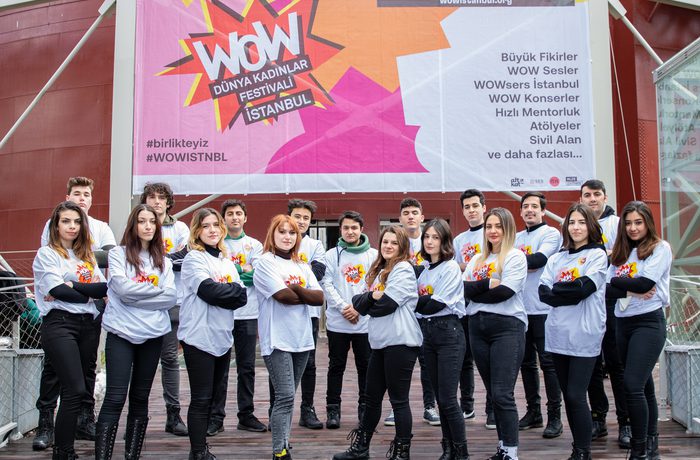 A group of young individuals standing confidently with crossed arms in front of a large, colourful "WOW Dünya Kadınlar Festivali İstanbul" banner