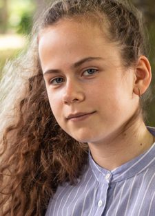 A young person with long curly hair stands by a tree, wearing a blue striped shirt