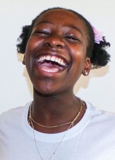 A young person with short curly hair and a flower accessory laughs joyfully. Wearing a white shirt and necklaces, they convey happiness against a plain background.