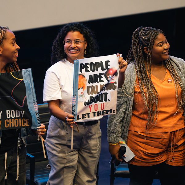 Three smiling women hold protest signs indoors. Signs read "My Body My Choice" and "People Are Talking About You. Let Them!" The atmosphere is empowering.