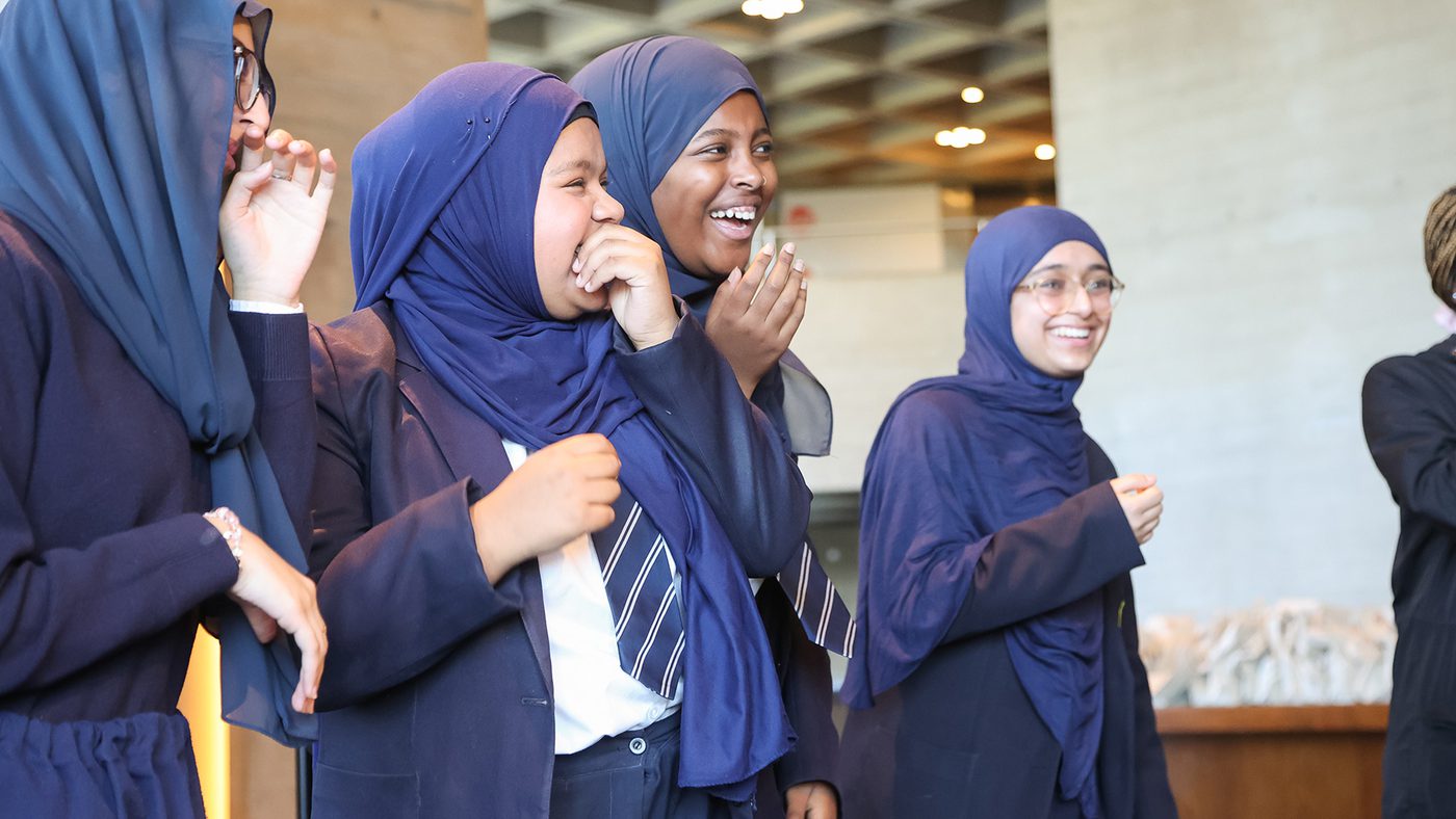 Four women wearing dark blue hijabs and uniforms laugh joyfully together indoors.