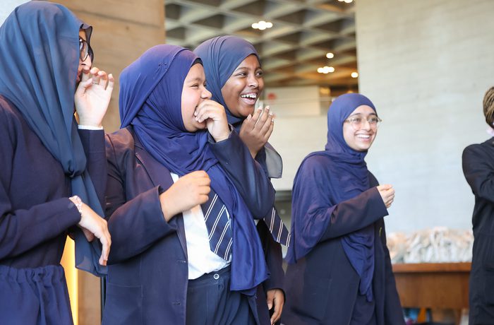 Four women wearing dark blue hijabs and uniforms laugh joyfully together indoors.