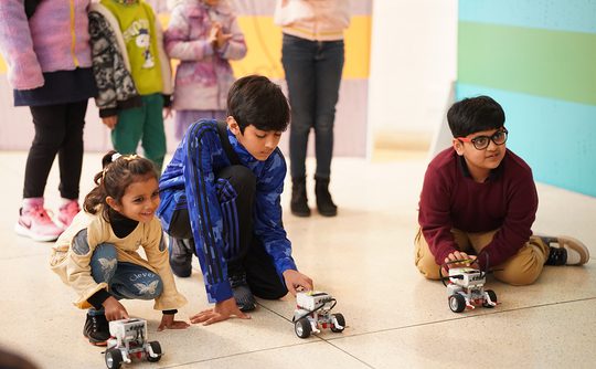 Children eagerly crouching with small robots on the floor, surrounded by spectators.