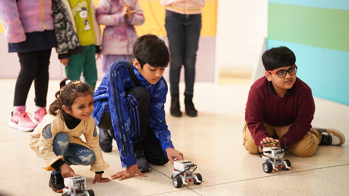 Children eagerly crouching with small robots on the floor, surrounded by spectators.