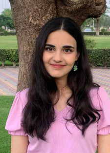 Young woman with long dark hair and a pink shirt stands smiling in a park.