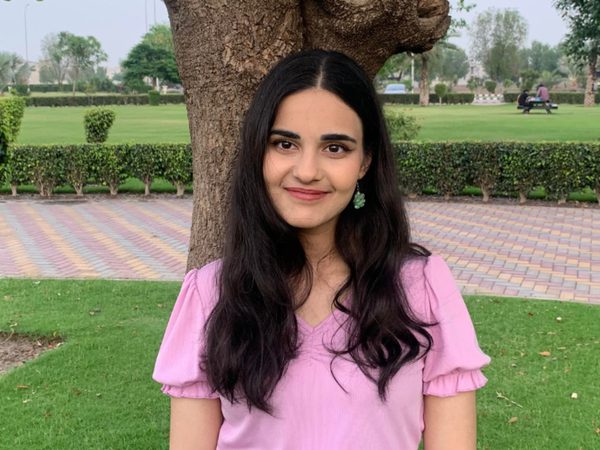 Young woman with long dark hair and a pink shirt stands smiling in a park.
